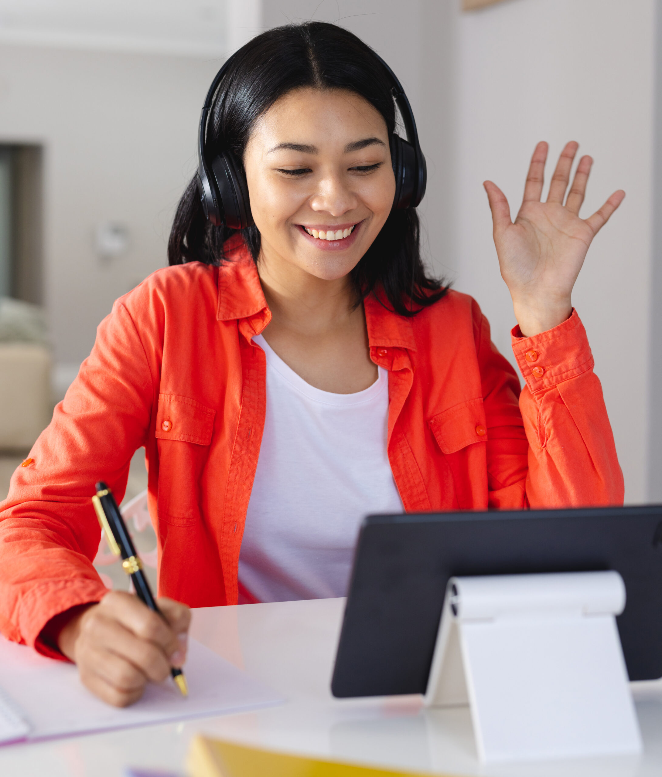 Vertical of happy biracial woman in headphones waving, making tablet video call at home, copy space. Working from home, technology, communication and lifestyle concept.